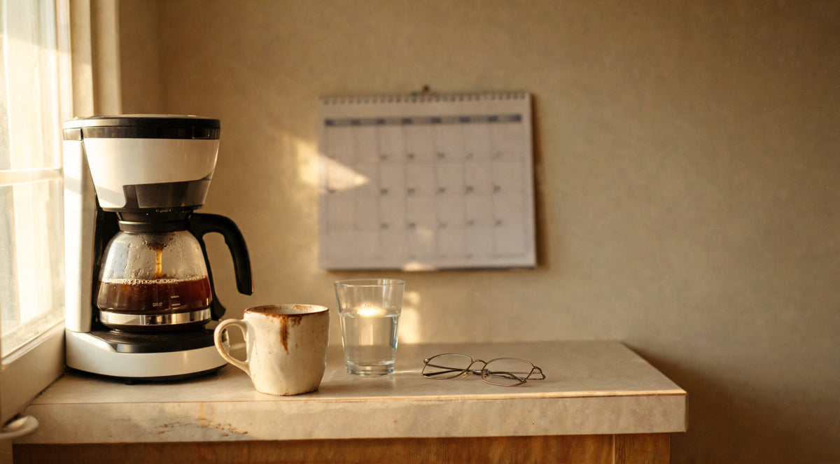 A coffee maker brewing in a kitchen corner, next to a used mug, a glass of water, and reading glasses on the counter below a wall calendar.