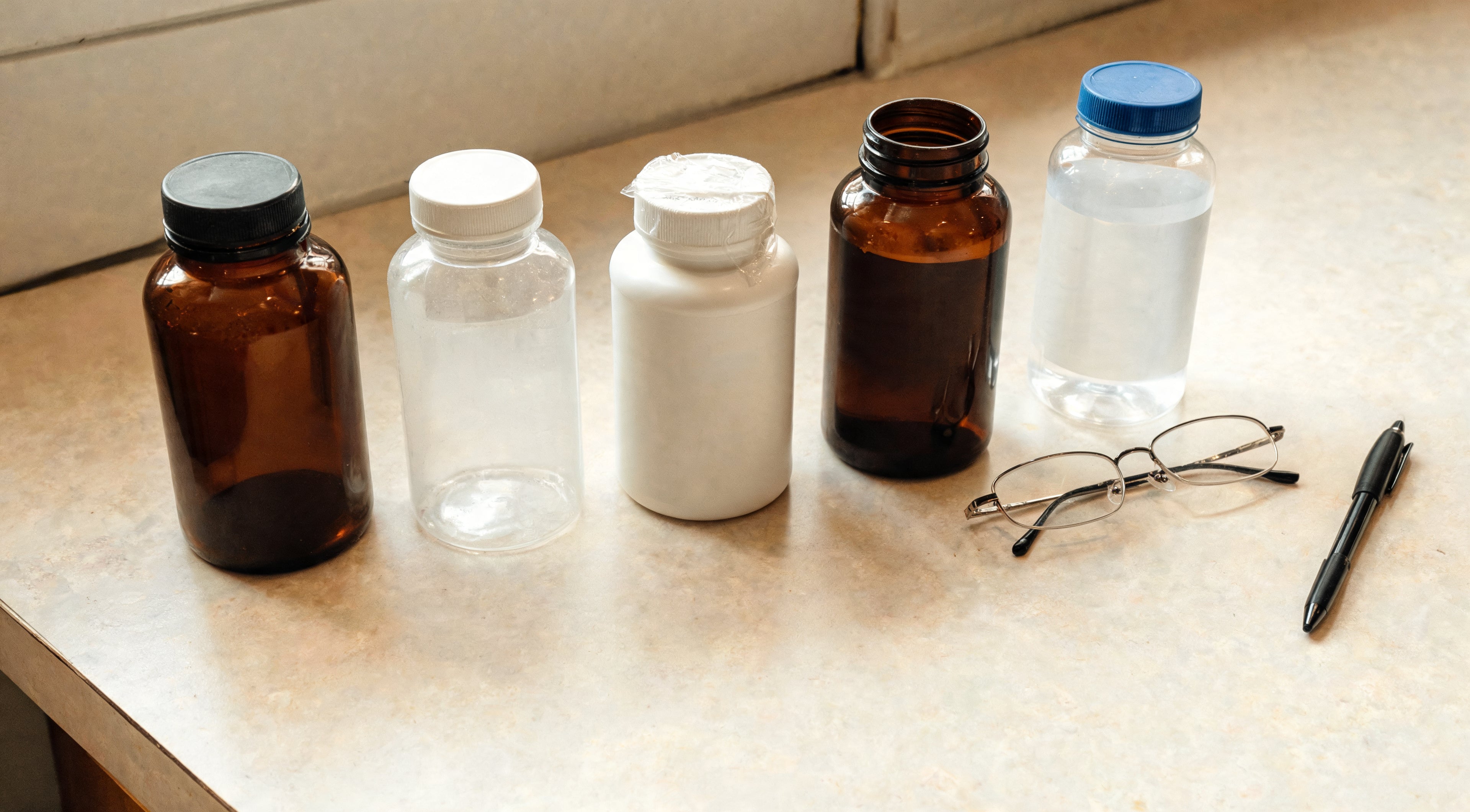 Five different medicine and supplement bottles lined up on a counter next to a pair of glasses and a pen.
