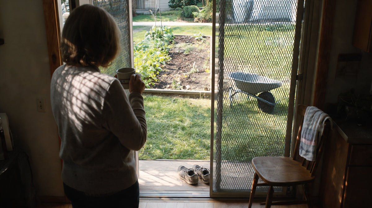 A woman stands inside a screen door holding a mug, looking out at a partly weeded garden bed.